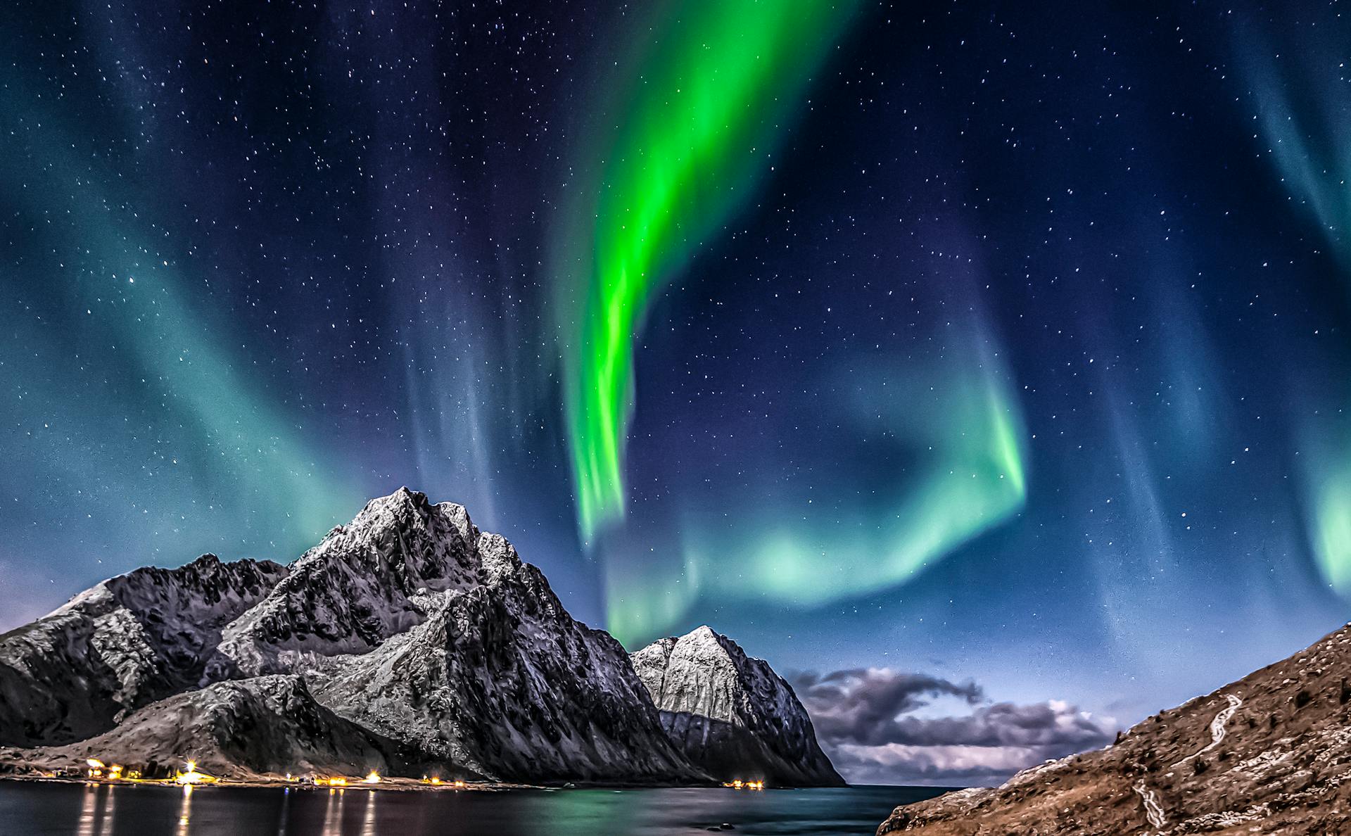 A large, snow-covered, icy mountain in the foreground with the northern lights in the sky above, with streaks of green, blue and white light.